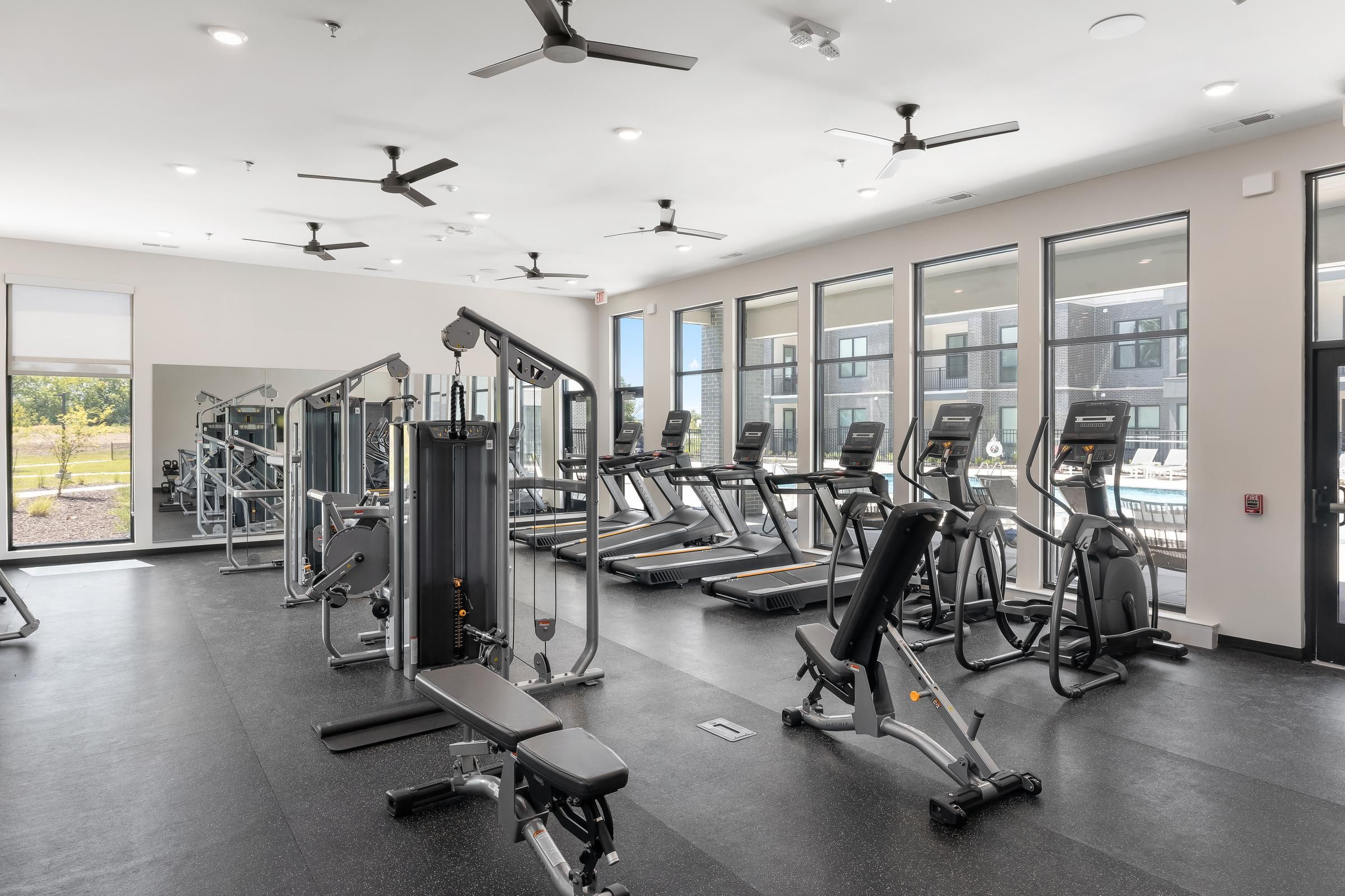 Modern gym with treadmills, weight machines, benches, and large windows letting in natural light.