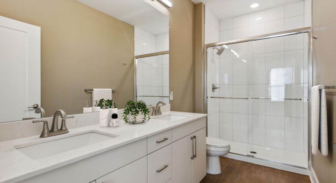 Modern bathroom with double sink, white cabinets, large mirror, glass shower, and wood-look flooring.