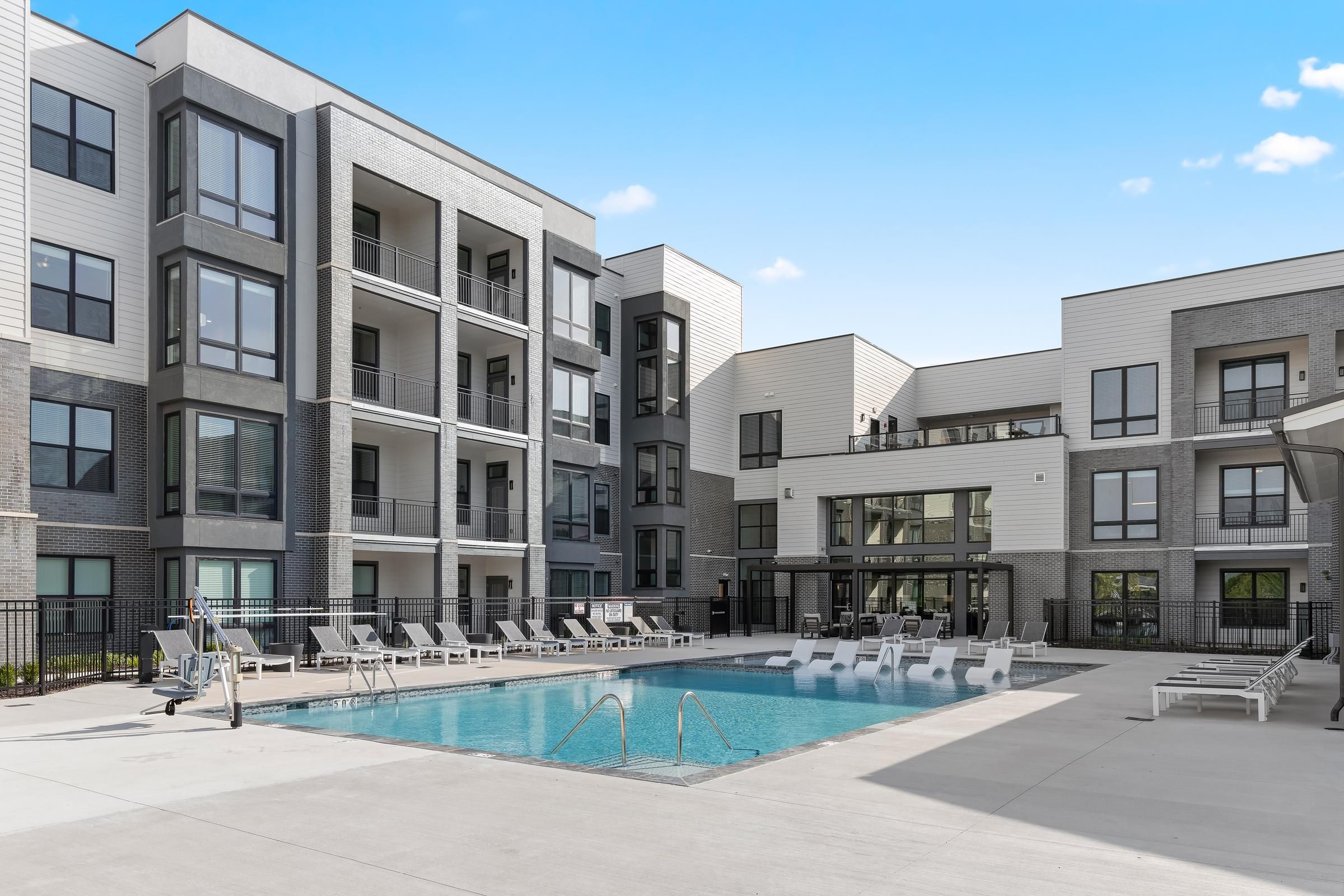 Modern apartment complex with balconies and a swimming pool surrounded by lounge chairs under a clear blue sky.