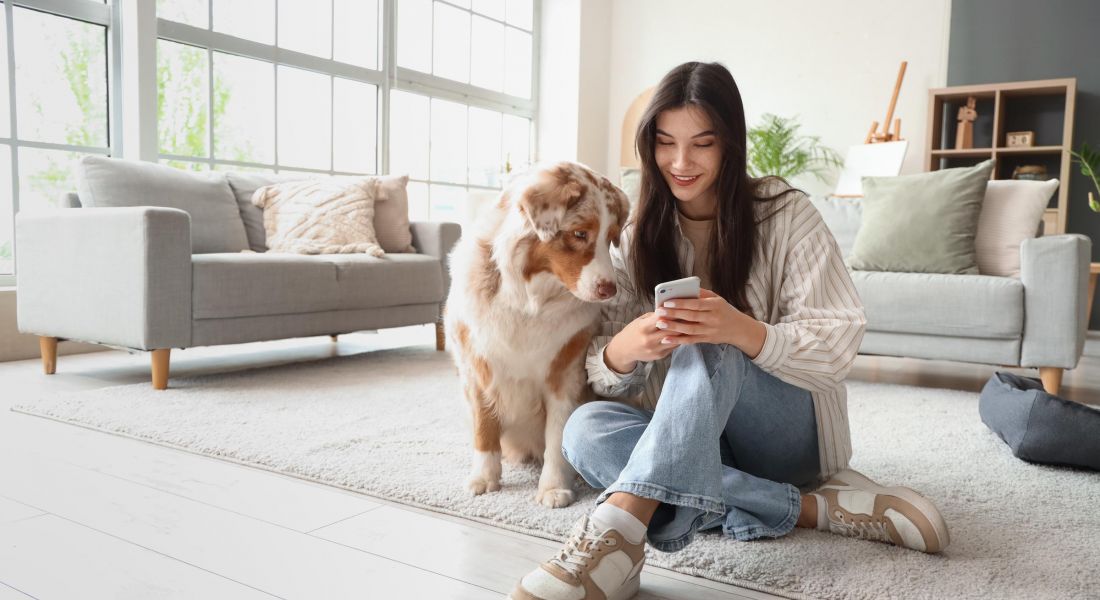 Woman sitting on the floor at home with a dog, smiling while looking at her phone.