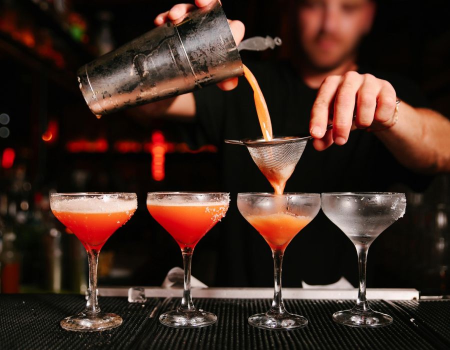 Bartender straining a red-orange cocktail into coupe glasses lined up on a bar counter.