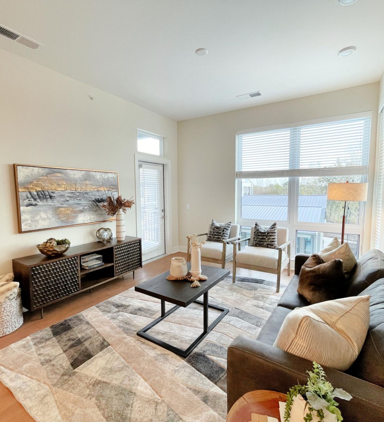 Modern living room with a dark sofa, large rug, wall art, two chairs by windows, and neutral decor.