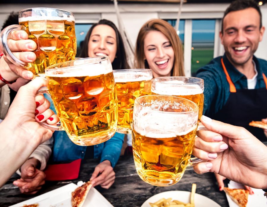 Friends clink beer mugs and smile while enjoying food outdoors in a lively, cheerful setting.
