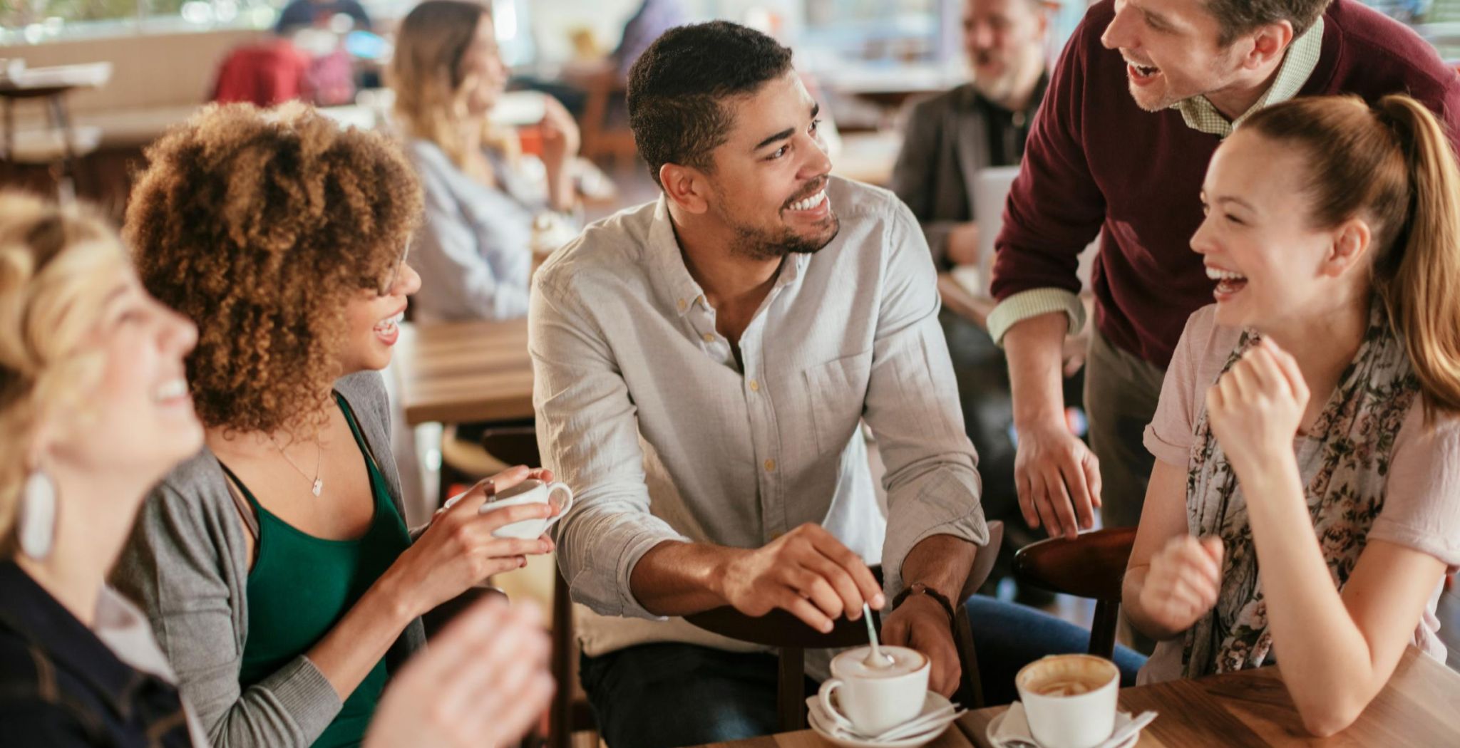 Group of friends laughing and chatting around a table with coffee at a bright, busy café.