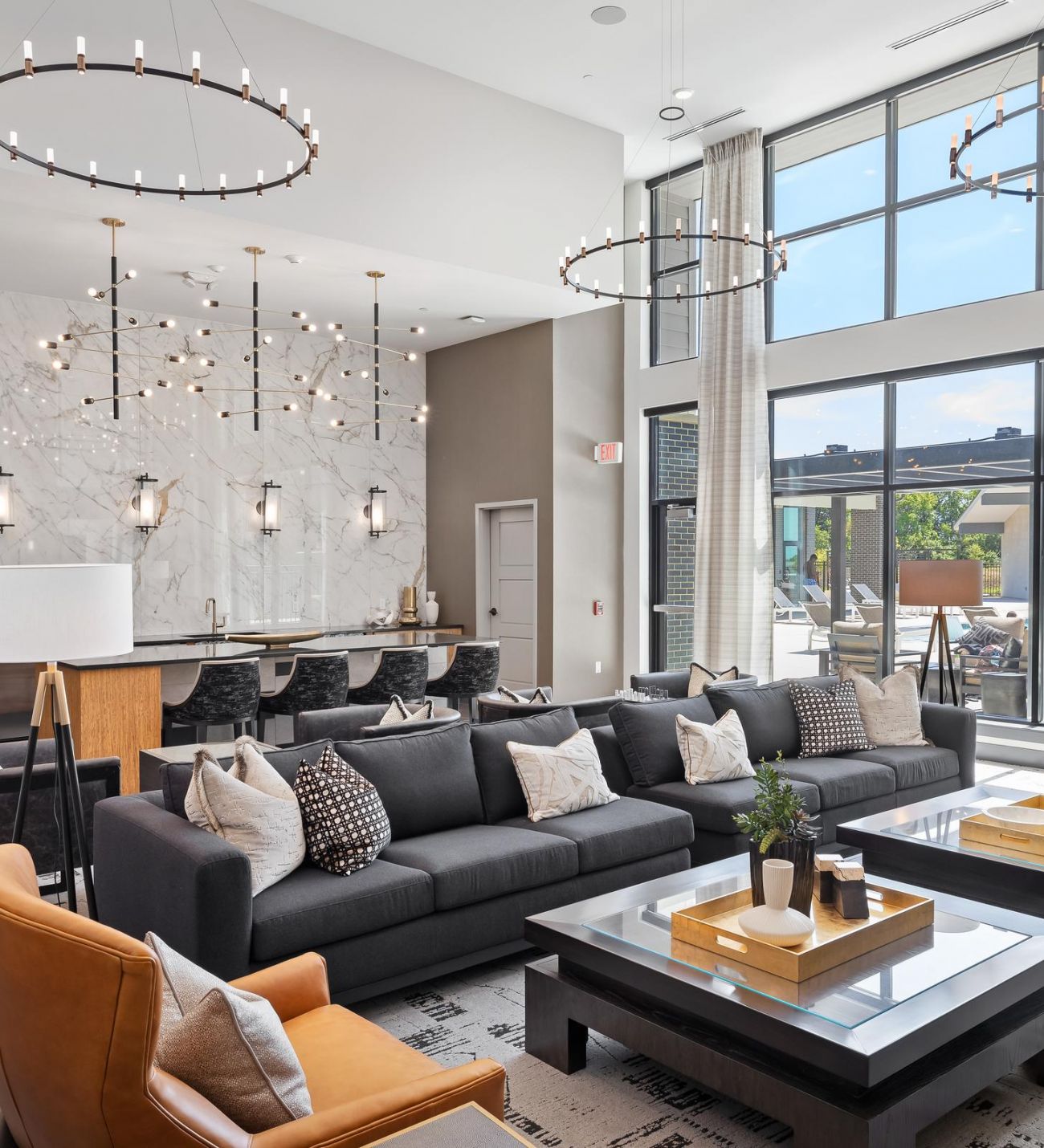 Modern lounge with sofas, armchairs, chandeliers, large windows, and a kitchen area in the background.