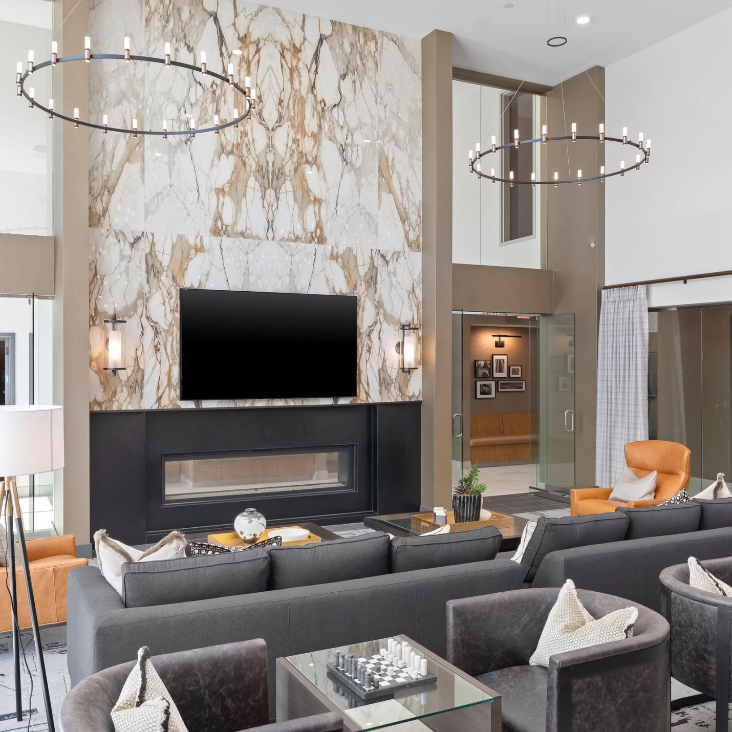 Modern lounge with gray sofas, armchairs, a marble fireplace, TV, and circular chandeliers overhead.