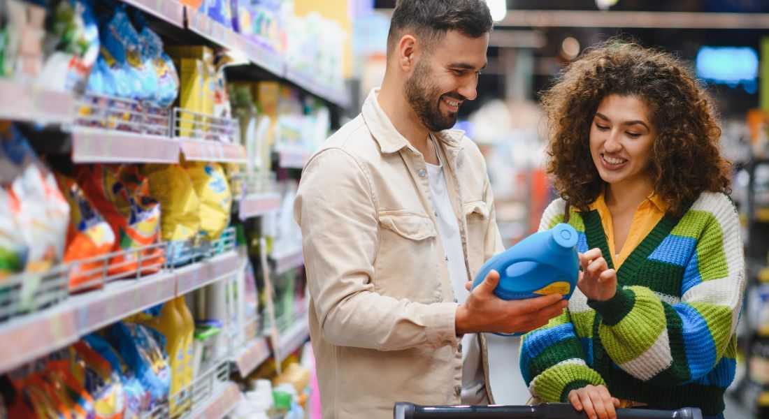 A man and woman smile while looking at a detergent bottle in a supermarket aisle.