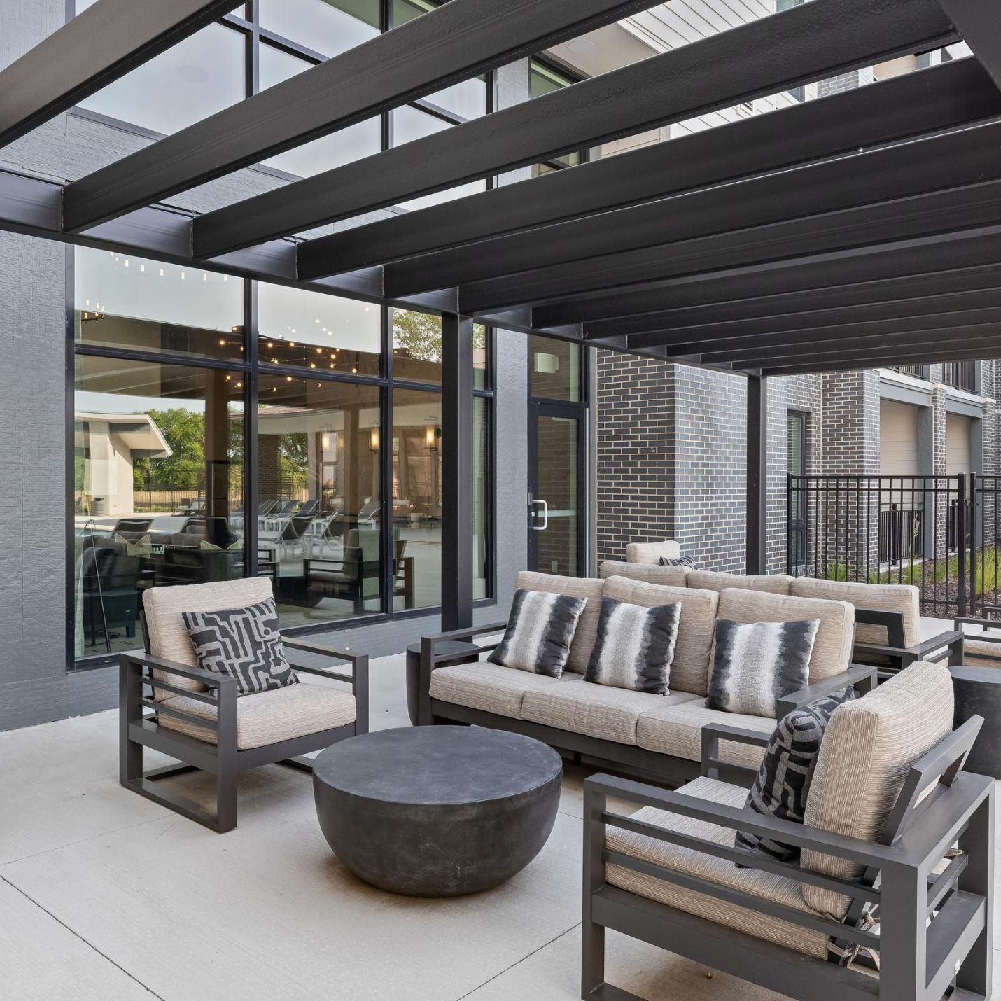 Modern outdoor patio with cushioned seating, a round coffee table, and a black pergola near a building.