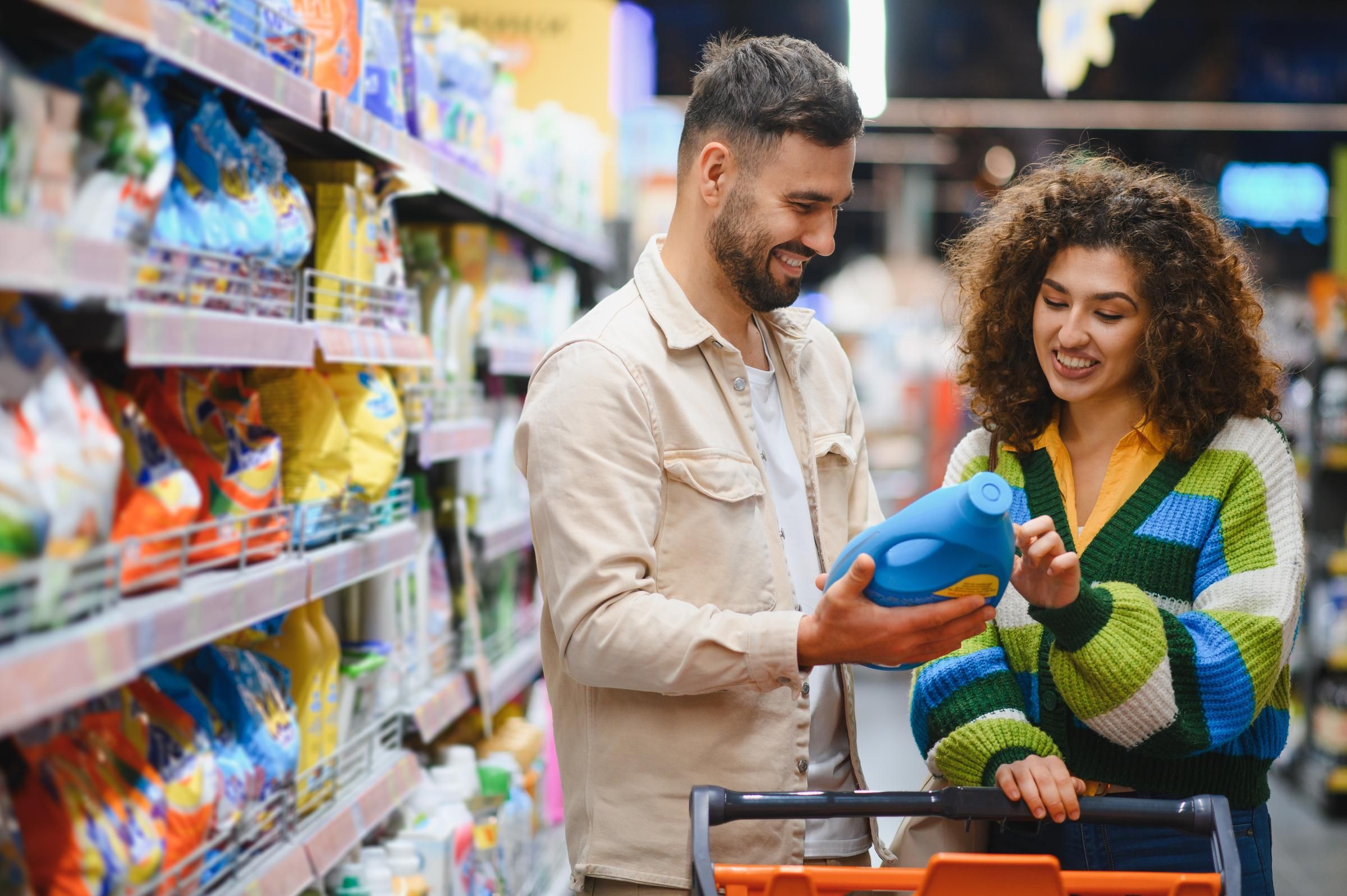 A man and woman smile while looking at a detergent bottle in a supermarket aisle.