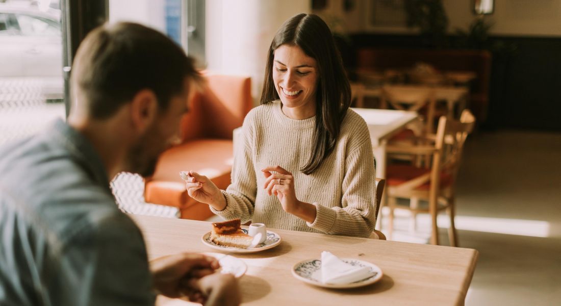 Two people sit at a café table, smiling and enjoying coffee and pastries in a bright, cozy setting.